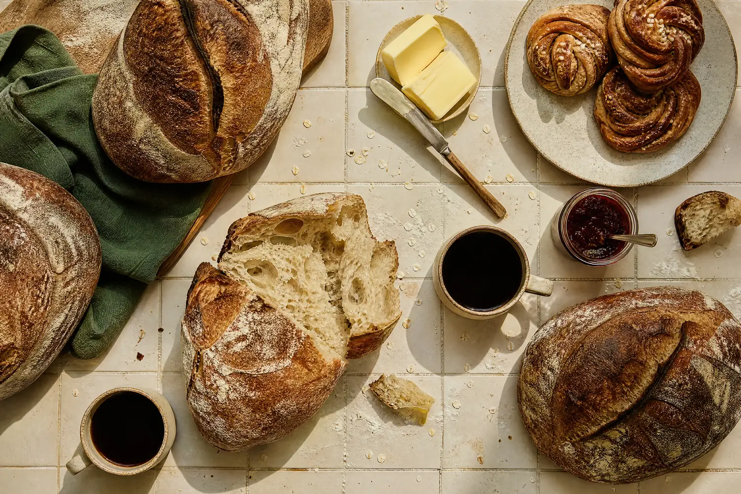 Ferske surdeigsbrød, kanelsnurrer, smør og kaffe på et flislagt bord